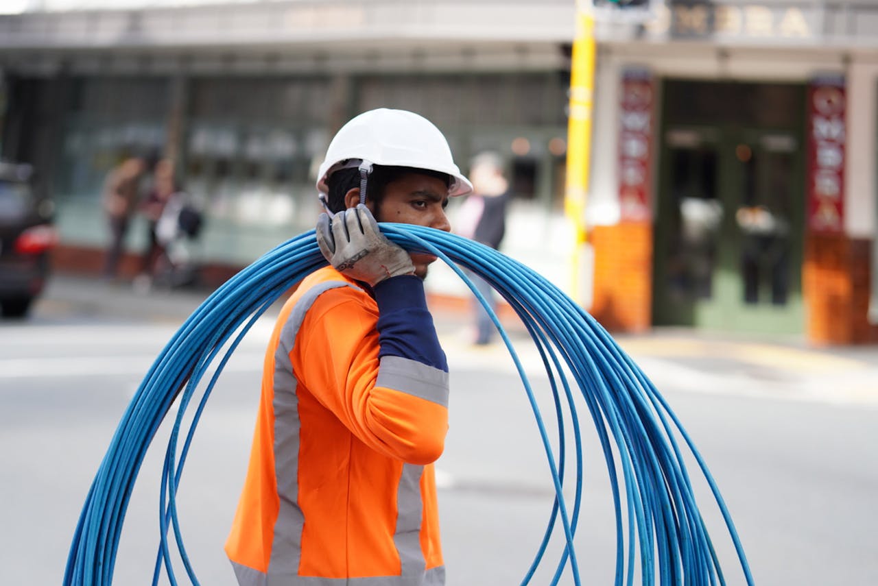 why-choose-us Construction worker in Wellington carrying blue cables, wearing safety gear on a city street.