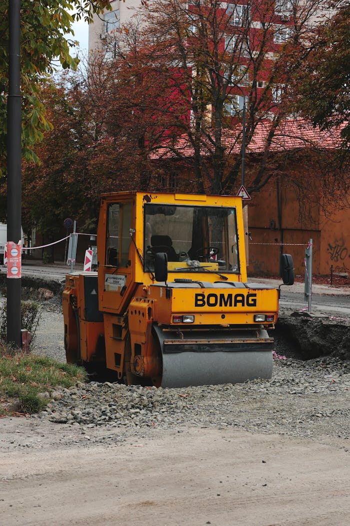team-04 Yellow BOMAG road roller at construction site with autumn trees in the background.